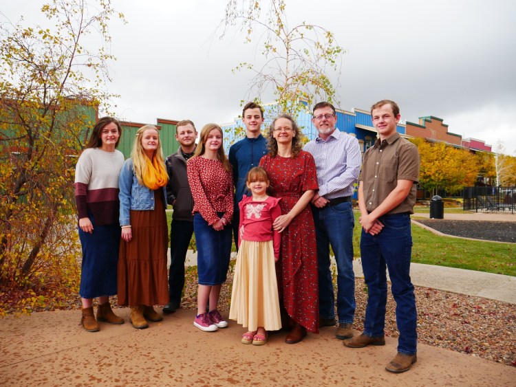 The Burton family, including Pastor Jason, his wife Amy, and their seven children, pose together outdoors in a casual family portrait, with colorful backgrounds and autumn foliage.